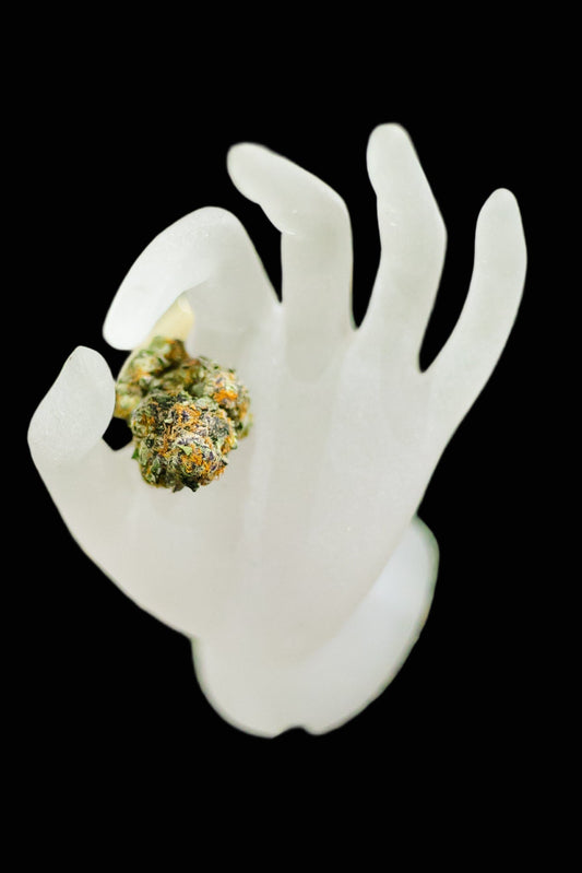 White ceramic hand holding a cannabis bud on a green surface with other cannabis items.
