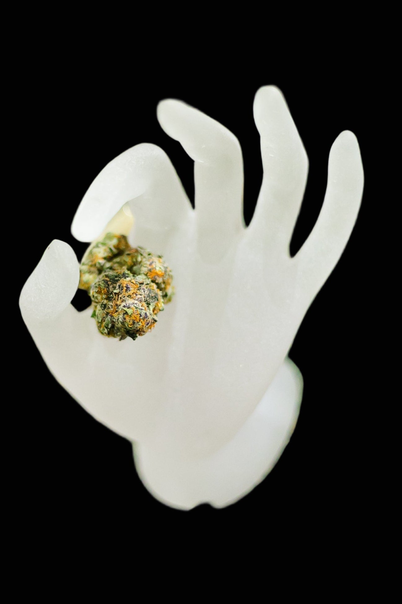 White ceramic hand holding a cannabis bud on a green surface with other cannabis items.